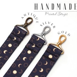 May include: Three camera straps with different colored clasps: antique brass, silver, and gold. Each strap is navy blue with a white moon and crescent moon pattern. The straps are handmade and have the text "Handmade Printed Straps" on the white background.