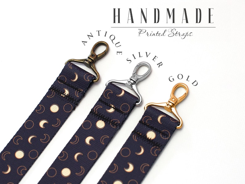 May include: Three camera straps with different colored clasps: antique brass, silver, and gold. Each strap is navy blue with a white moon and crescent moon pattern. The straps are handmade and have the text "Handmade Printed Straps" on the white background.