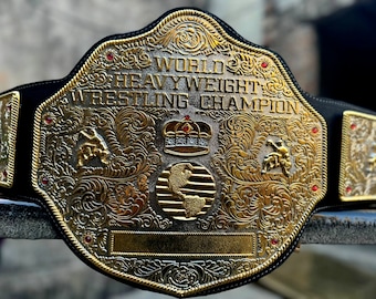 May include: A gold-colored championship belt with a black leather strap. The belt features a large, ornate center plate with the words "World Heavyweight Wrestling Champion" and a crown design. The belt is decorated with red jewels.