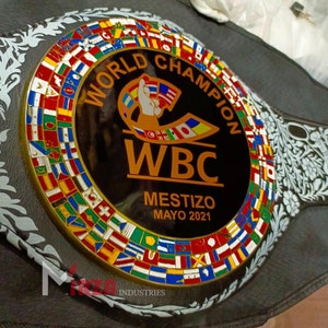May include: A black and gold boxing championship belt with a black and white floral design. The belt features a circular emblem with the text "World Champion WBC Mestizo Mayo 2021" and a ring of flags representing different countries.