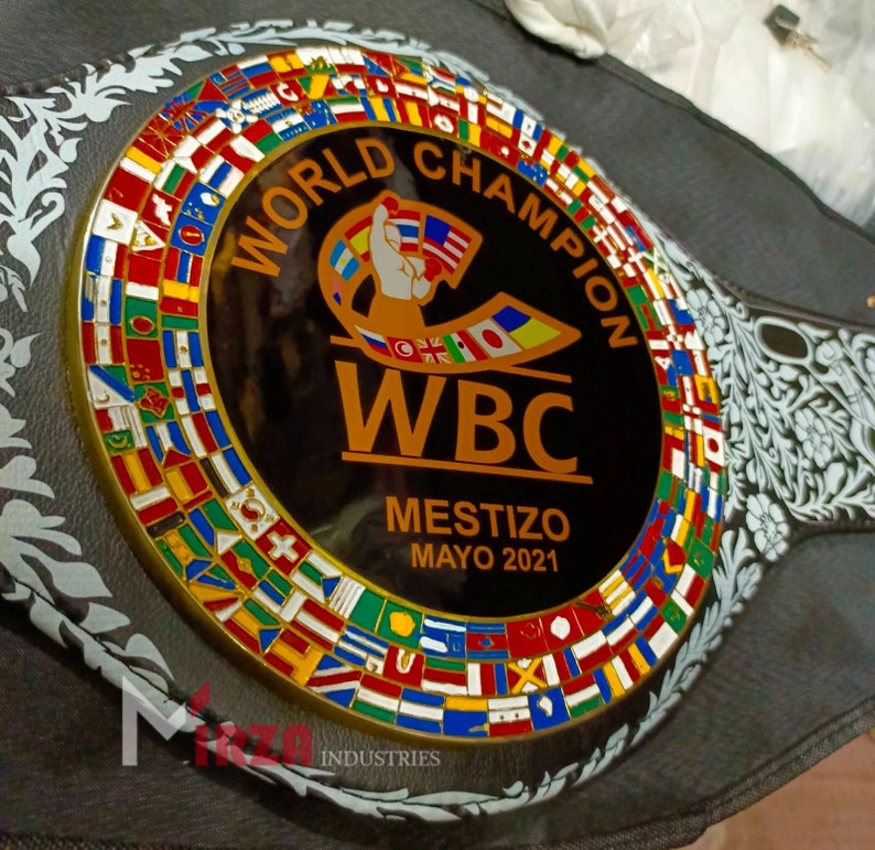 May include: A black and gold boxing championship belt with a black and white floral design. The belt features a circular emblem with the text "World Champion WBC Mestizo Mayo 2021" and a ring of flags representing different countries.