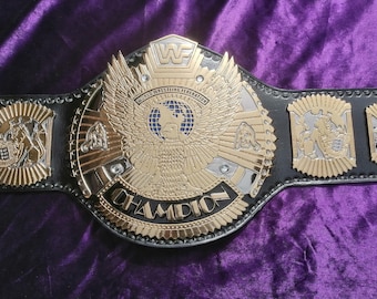 Puede incluir: Un cinturón de campeonato de lucha libre dorado y plateado con una correa de cuero negro. La placa central presenta un águila grande con las palabras "World Wrestling Federation" y "Champion". Las placas laterales tienen diseños dorados y plateados.