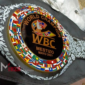 May include: A black leather boxing championship belt with a gold-coloured buckle featuring a circle of flags from around the world. The centre of the buckle has a black background with the words "World Champion" in gold lettering, a gold WBC logo, and the text "Mestizo Mayo 2021".