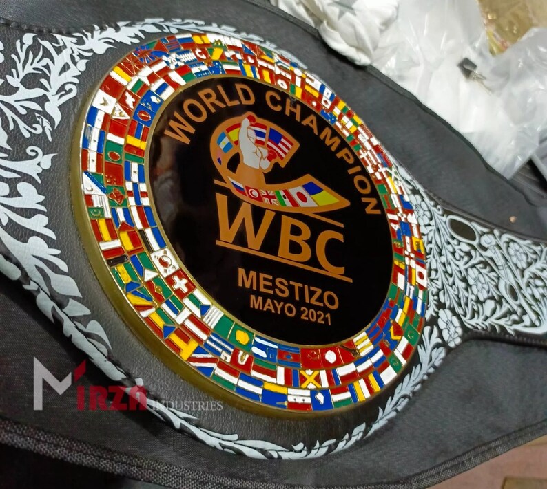 May include: A black leather boxing championship belt with a gold-coloured buckle featuring a circle of flags from around the world. The centre of the buckle has a black background with the words "World Champion" in gold lettering, a gold WBC logo, and the text "Mestizo Mayo 2021".