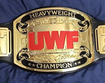 Puede incluir: Un cinturón de campeonato de lucha libre dorado y negro con las palabras "Heavyweight" y "Universal Wrestling Federation" en la parte delantera. El cinturón también tiene las palabras "UWF Champion" en la parte delantera.