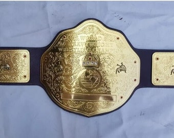 May include: A gold-colored championship belt with a large, ornate center plate that reads "World Heavyweight Wrestling Champion". The belt has a brown leather strap with gold-colored side plates.