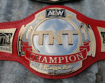 May include: A red and gold wrestling championship belt with the text "TNT CHAMPION" on a red banner. The center features the letters "TNT" in gold, with "AEW WRESTLING" above. The belt has intricate gold detailing.