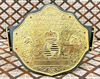 May include: A gold-colored wrestling championship belt with a black leather strap. The belt features a large, ornate center plate with the words "World Heavyweight Wrestling Champion" and a globe design. The belt is decorated with red jewels and intricate engravings.