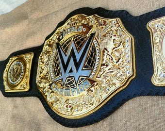 May include: A gold and black championship belt featuring the WWE logo. The belt is highly detailed, with the word "CHAMPION" and ornate designs. It is made of gold-coloured metal and black leather, with a total length of approximately 120 cm.