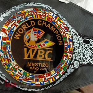 May include: A black and gold boxing championship belt with the words "World Champion" and "WBC" on it. The belt is decorated with a circle of flags from different countries and a floral design.