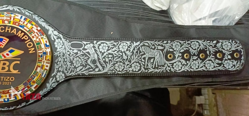 May include: A black leather boxing championship belt with gold accents and a large circular centre plate featuring the text "WBC Champion" and a colourful design of flags. The belt has a silver floral design with a horse and rider.