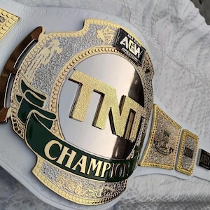 May include: A championship belt with a white leather strap and gold and silver accents. The center plate features the letters "TNT" in gold, with "CHAMPION" in gold lettering below. The belt also has the text "AEW ELITE WRESTLING".