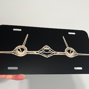 May include: Black license plate with a beige graphic design. The design features a stylized symmetrical image with two circular elements and a central geometric shape. The plate has four mounting holes.
