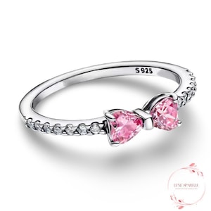 May include: A silver ring with a bow design. The bow features two pink heart-shaped stones. The ring band is adorned with small, clear stones. The ring is marked with "S925".