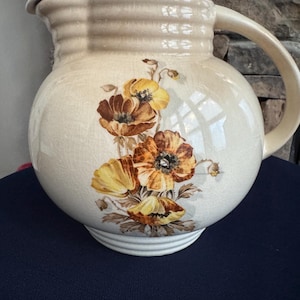 May include: Cream-colored ceramic pitcher with a lid and handle. The pitcher features a floral design with yellow and brown flowers. The pitcher has a round body and a slightly textured surface. A vintage-style kitchenware item.