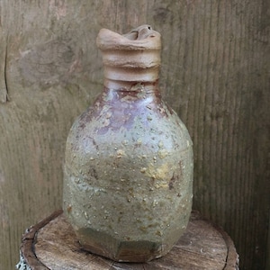 以下が含まれることがあります： 茶色、緑、金色の斑点のある釉薬をかけた、素朴な手作りの陶器の花瓶。花瓶は、幅広で丸みを帯びた底と、細い首をしています。