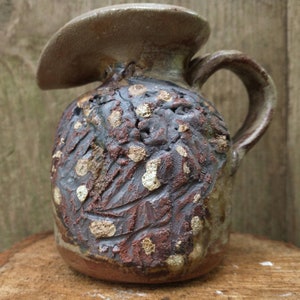 May include: A small, brown and gray ceramic pitcher with a wide, flat spout and a handle. The pitcher has a textured surface with white and brown spots.