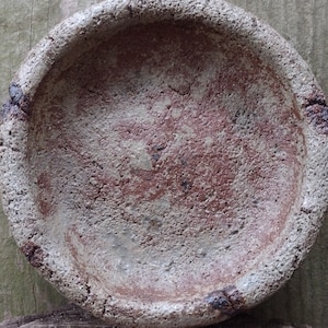 Op de afbeelding: Een klein, rond, grijs keramisch schaaltje met een ruw, getextureerd oppervlak. Het schaaltje heeft een roodbruin interieur en een licht verhoogde rand.