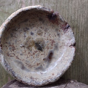 May include: A small, round, ceramic dish with a speckled, brown and white glaze. The dish has a slightly uneven rim and a rough, textured surface.