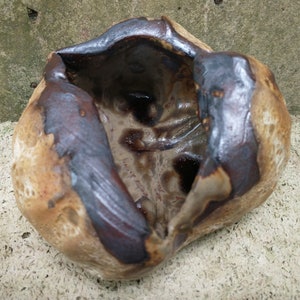 Op de afbeelding: Een handgemaakte keramische kom met een unieke, organische vorm. De kom heeft een mix van bruine en donkerbruine kleuren, met een glanzend, getextureerd interieur. De buitenkant heeft een ruwe, natuurlijke afwerking, die het vakmanschap laat zien.