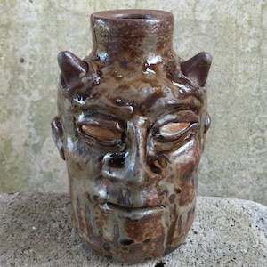 May include: A handcrafted ceramic vase, featuring a face design. The vase is predominantly brown, with darker brown and grey accents. It has a detailed face with closed eyes, a small mouth, and two small horns. The vase has a cylindrical shape with a wide opening.