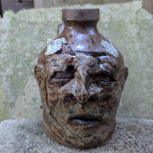May include: A brown ceramic face jug with a narrow neck and a handle. The vessel features a sculpted human face with deep-set eyes, a prominent nose, and an open mouth. The surface has a mottled, glossy glaze with textured white patches.