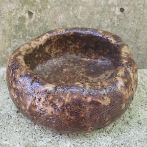 Op de afbeelding: Een kleine, ronde, bruine keramische pot met een ruw, getextureerd oppervlak. De pot heeft een brede, ondiepe opening en is perfect voor kleine planten of vetplanten.