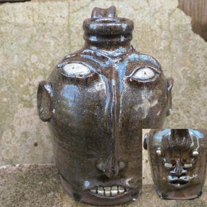 May include: A handmade, brown ceramic jug with a face design. It has white eyes, a large nose, and a smiling mouth with teeth. The jug has a handle-like feature on top and ear-like shapes on the sides. The jug is approximately 20 cm tall.