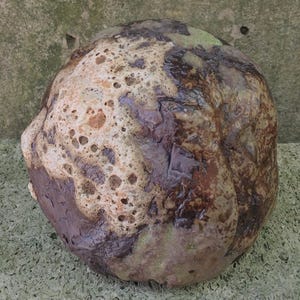 Op de afbeelding: Een ronde, bruine en witte keramische sculptuur met een getextureerd oppervlak. De sculptuur heeft een ruwe, ongelijke textuur en lijkt van klei te zijn gemaakt.