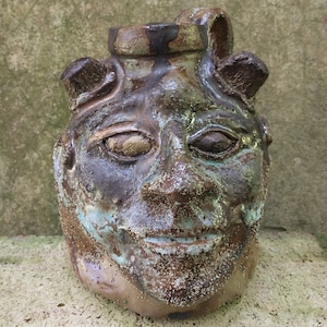 May include: A ceramic jug with a face sculpted on the front. The jug is brown, black, and blue with a textured surface.
