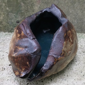 Puede incluir: Una pequeña maceta de cerámica marrón oscuro con una superficie rugosa y texturizada. La maceta está abierta en la parte superior y tiene un esmalte azul oscuro en el interior.