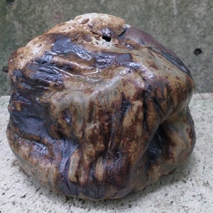 Op de afbeelding: Een kleine, onregelmatig gevormde keramische pot met een bruine, zwarte en witte glazuur. De pot heeft een ruw, getextureerd oppervlak en lijkt handgemaakt te zijn.