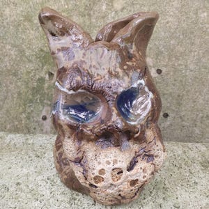 以下が含まれることがあります： 青い目をした猫の頭のセラミック彫刻。彫刻は茶色で、ざらざらとした質感の表面をしています。