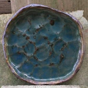 Op de afbeelding: Een tealblauwe keramische schaal met een getextureerd, onregelmatig oppervlak. De schaal heeft een bruine rand en is licht onregelmatig van vorm.