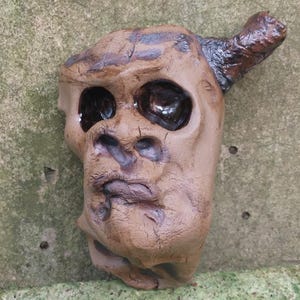 Op de afbeelding: Een bruine keramische sculptuur van een gezicht met een enkele hoorn. Het gezicht heeft diepliggende ogen en een getextureerd oppervlak.