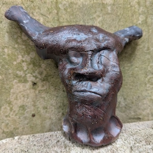Op de afbeelding: Een handgemaakte, abstracte sculptuur van een gezicht met hoorns, in tinten bruin en grijs. De sculptuur heeft een getextureerd oppervlak en een enigszins verweerde uitstraling, met zichtbare scheuren die bijdragen aan het karakter. Het gezicht heeft een strenge uitdrukking.