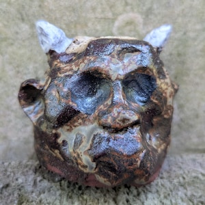 Op de afbeelding: Handgemaakte keramische sculptuur van een gezicht met hoorns en grote oogkassen. Het gezicht is voornamelijk bruin met lichtere accenten en een roodachtige basis. De hoorns zijn wit. De sculptuur heeft een ruwe, getextureerde uitstraling.