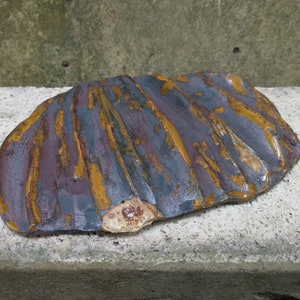 Pode incluir: Uma pedra decorativa grande, de forma irregular, com uma superfície texturizada. A pedra apresenta uma paleta de cores roxo escuro, cinza e amarelo dourado, com uma área mais clara de cor bege em um lado. O design da pedra tem um padrão canelado.