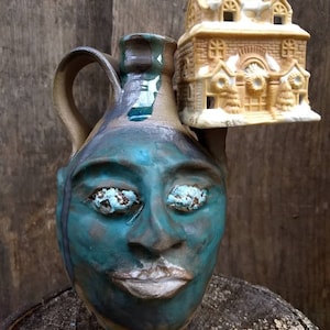 May include: A blue ceramic jug with a face and closed eyes, topped with a small ceramic house with white snow accents.