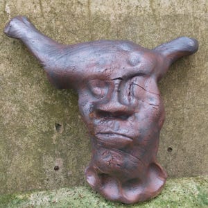 May include: A brown ceramic sculpture of a face with a long, thin nose and a wide mouth. The sculpture is textured and has a rough, uneven surface.