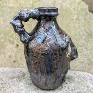 May include: A handmade ceramic jug with a dark, textured glaze. The jug features a handle and a spout, with a mix of brown, black, and metallic accents. The rustic pottery has a unique, organic shape.