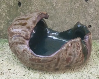 Puede incluir: Un pequeño cuenco de cerámica con un exterior texturizado marrón y un interior liso verde oscuro. El cuenco tiene forma de concha y un diseño orgánico único.