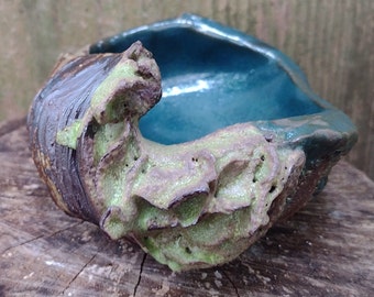 May include: A ceramic planter with a textured, green and brown exterior. The planter has a teal blue interior and a unique, organic shape.