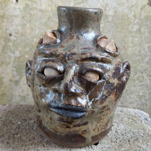 May include: A handcrafted ceramic vase, featuring a face design. The vase is predominantly brown and grey, with a textured finish. The face has stone eyes and a dark mouth. A unique art piece.
