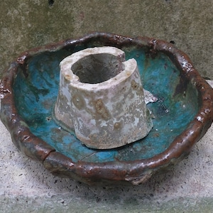 May include: A teal blue ceramic dish with a brown rim and a white ceramic cylinder in the center. The cylinder has a hole in the top and a rough, textured surface.