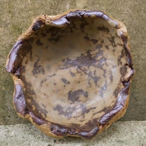 Op de afbeelding: Een bruine keramische kom met een getextureerde, ongelijke rand. De kom heeft een gevlekt bruin en tan glazuur met een licht iriserende afwerking.