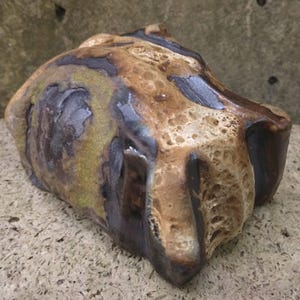 Op de afbeelding: Een keramisch beeldhouwwerk met een ruw, getextureerd oppervlak. Het beeldhouwwerk is bruin, beige en grijs met een gevlekt, abstract patroon.