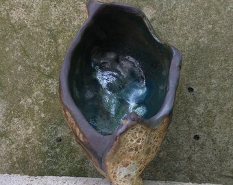 May include: A small, ceramic bowl with a rough, textured exterior and a smooth, dark blue interior. The bowl is shaped like a shell and has a unique, organic form.