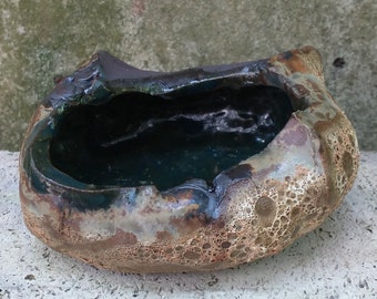 May include: A small, irregularly shaped ceramic planter with a dark green glaze inside and a brown, speckled glaze on the outside. The planter has a rough, textured surface.