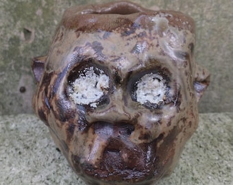 May include: A brown ceramic planter shaped like a face with white glaze accents for the eyes. The planter has a rough, textured surface.